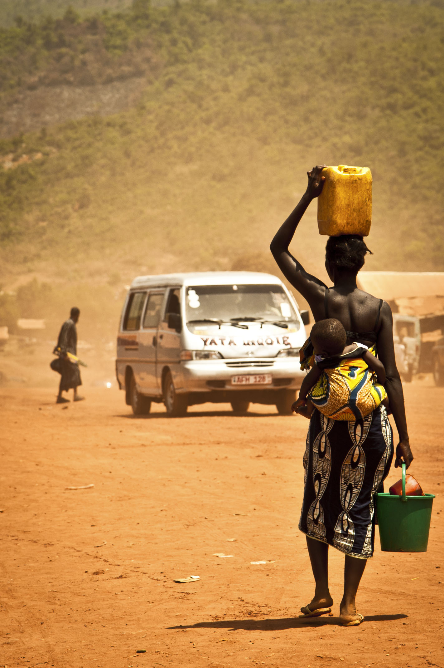 A woman carrying an infant and water containers walks down a dirt road. Photo by Eduardo Arraes