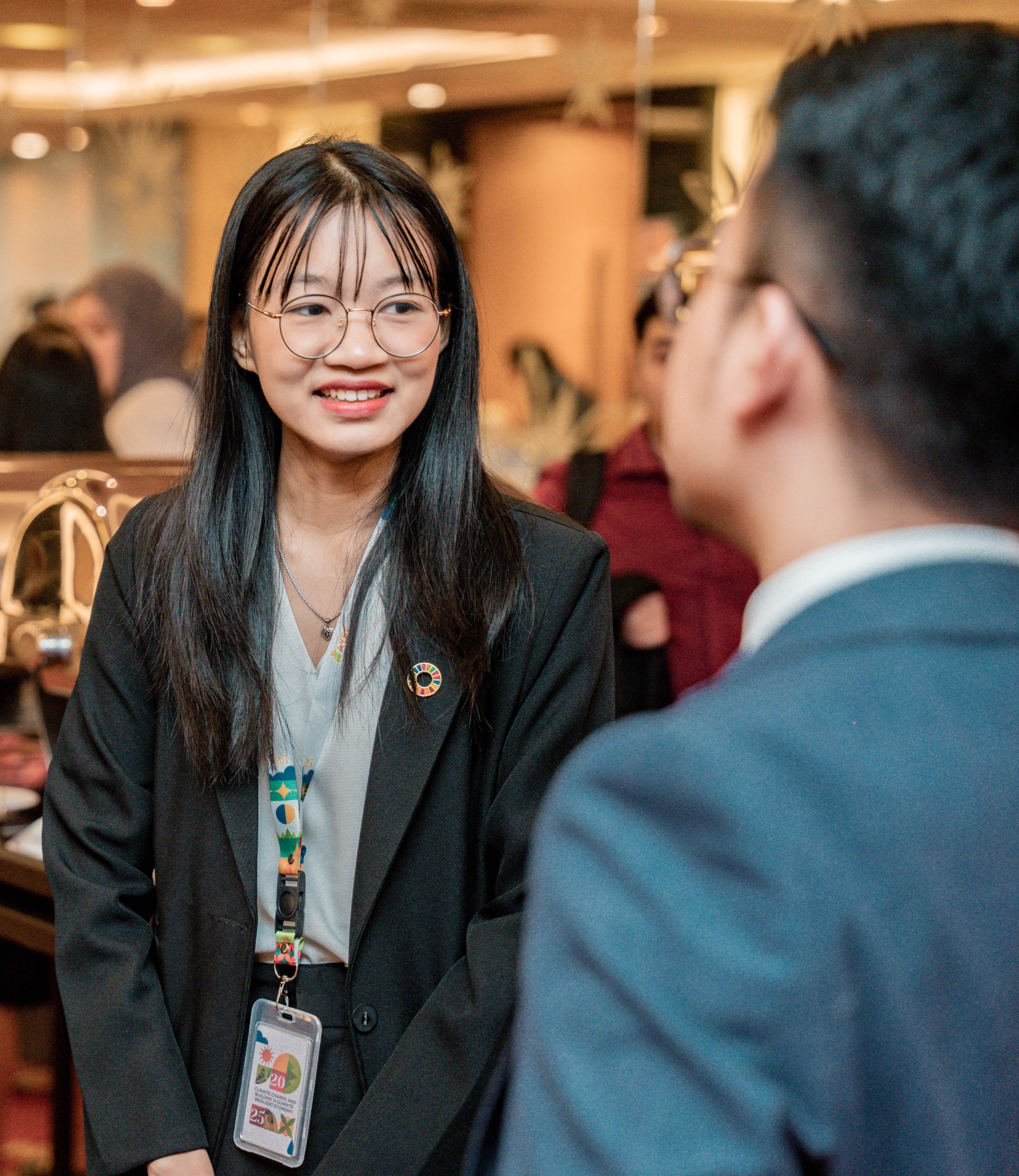 A well-dressed young woman sporting glasses and a a badge speaks to someone else in a lobby.