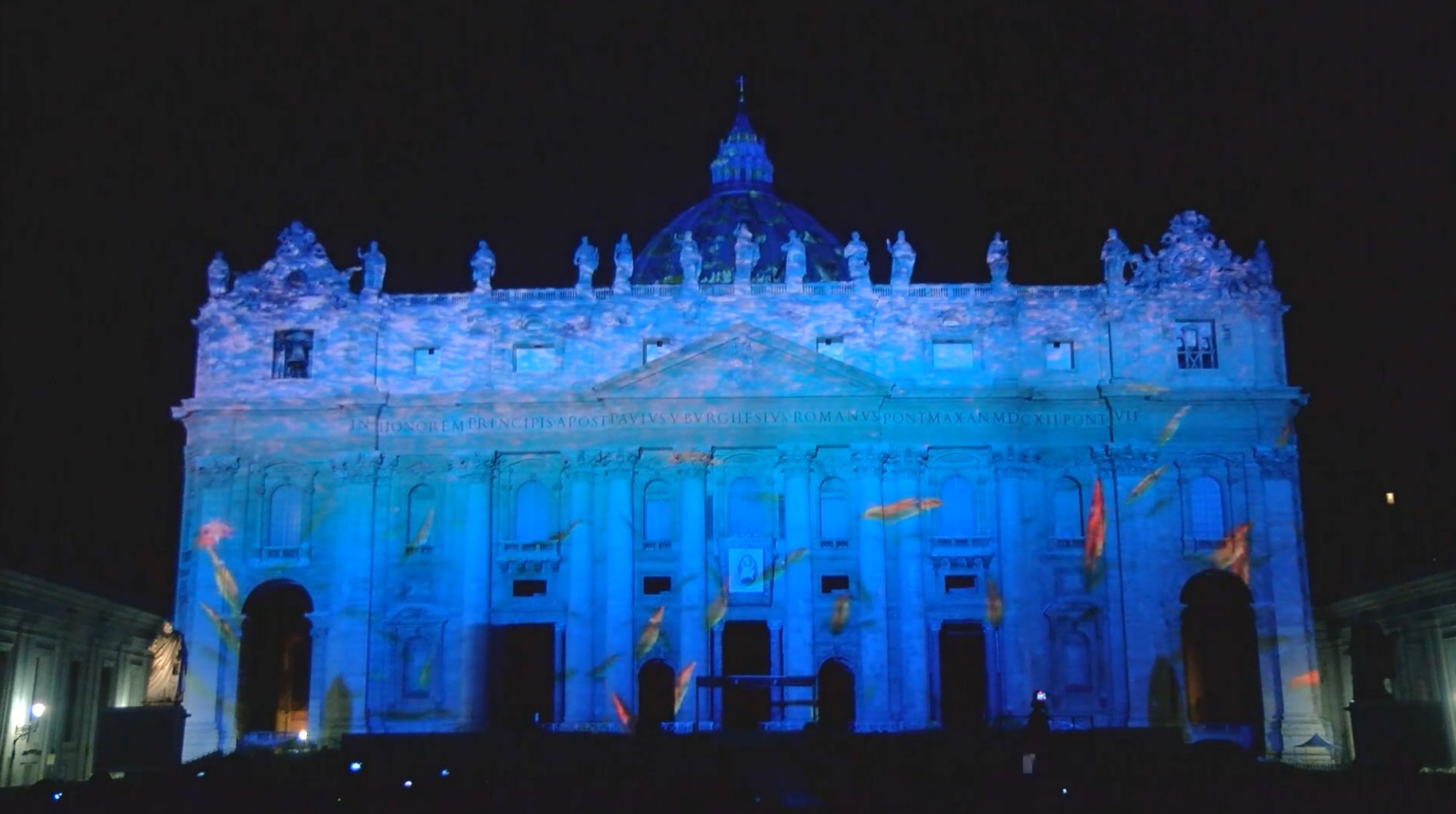 The facade of St. Peter's Basilica awash in blue light as an ocean scene is projected on it.