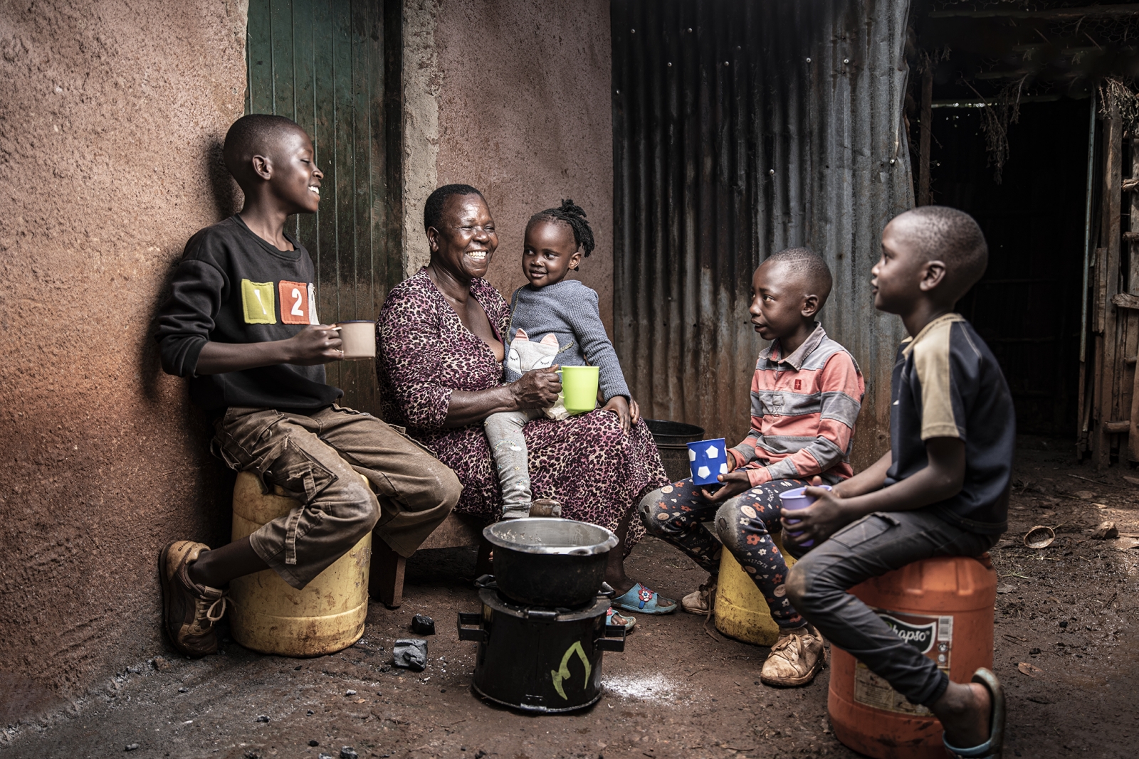 A Kenyan family sits around a stove with mugs in hand.