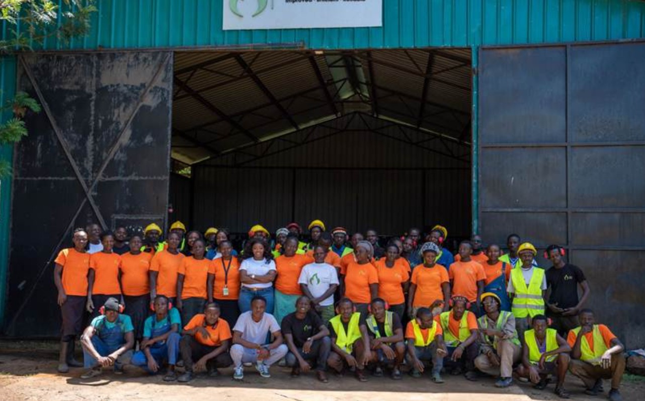 Several dozen employees, some in orange shirts and others in high-vis vests, pose for a group photo outside a large warehouse.