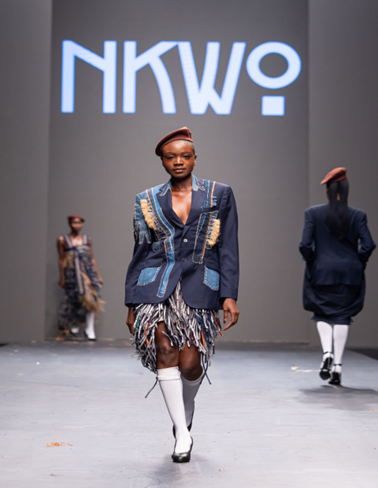 An African model in a rakish tan cap, a blue and gold jacket, a silvery fringe skirt, and long white socks.