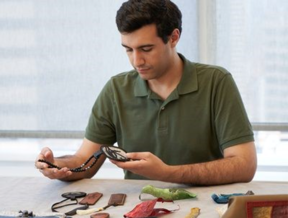 A young man in a forest-green polo handles a beaded necklace depicting the eye of Ra.