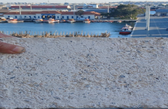 Background: ships moored in a harbor. Foreground: a hand holding up a textured building material resembling a cobbled street.