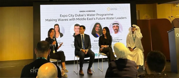 A young woman with a mic speaks onstage alongside three other young people. A projected slide in the background reads: "Expo City Dubai's Water Programme Making Waves with Middle East's Future Water Leaders."