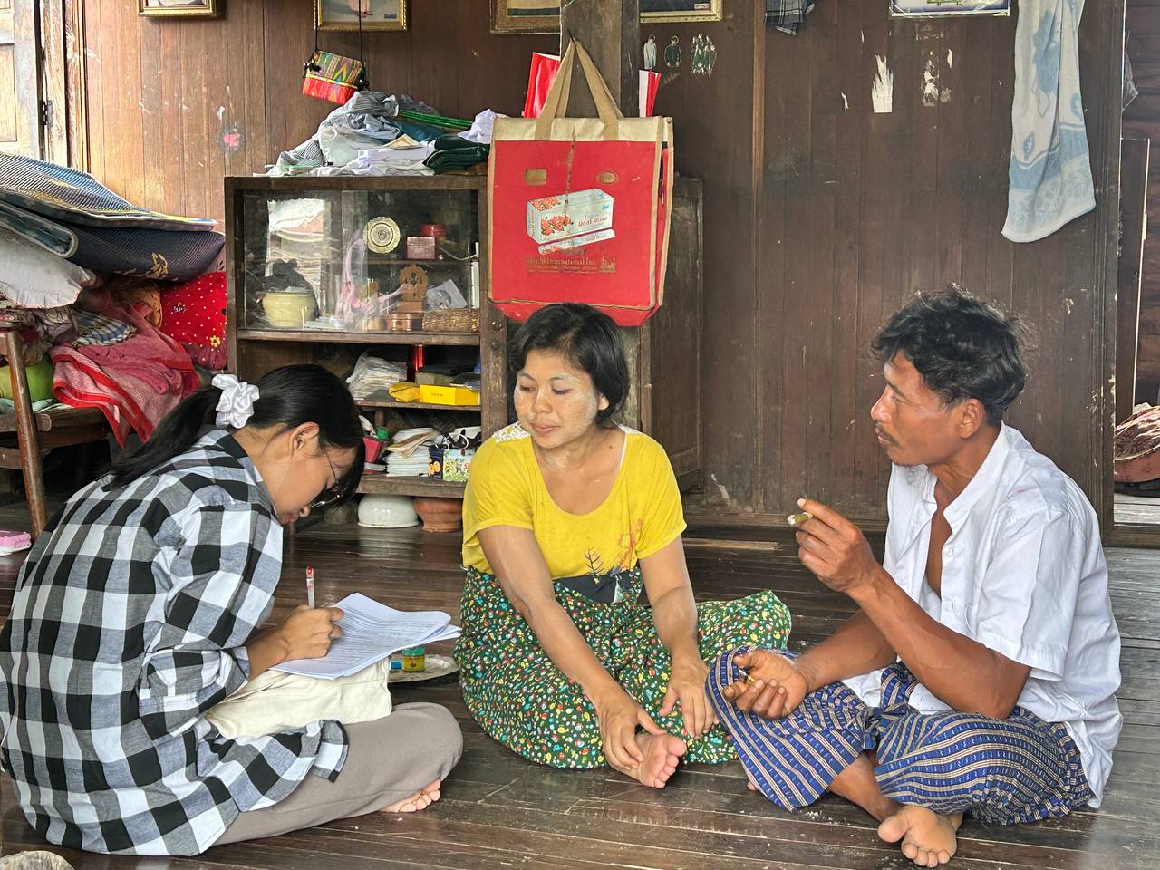 A young woman sits cross-legged on the floor with an older couple, collecting survey data and writing it down.