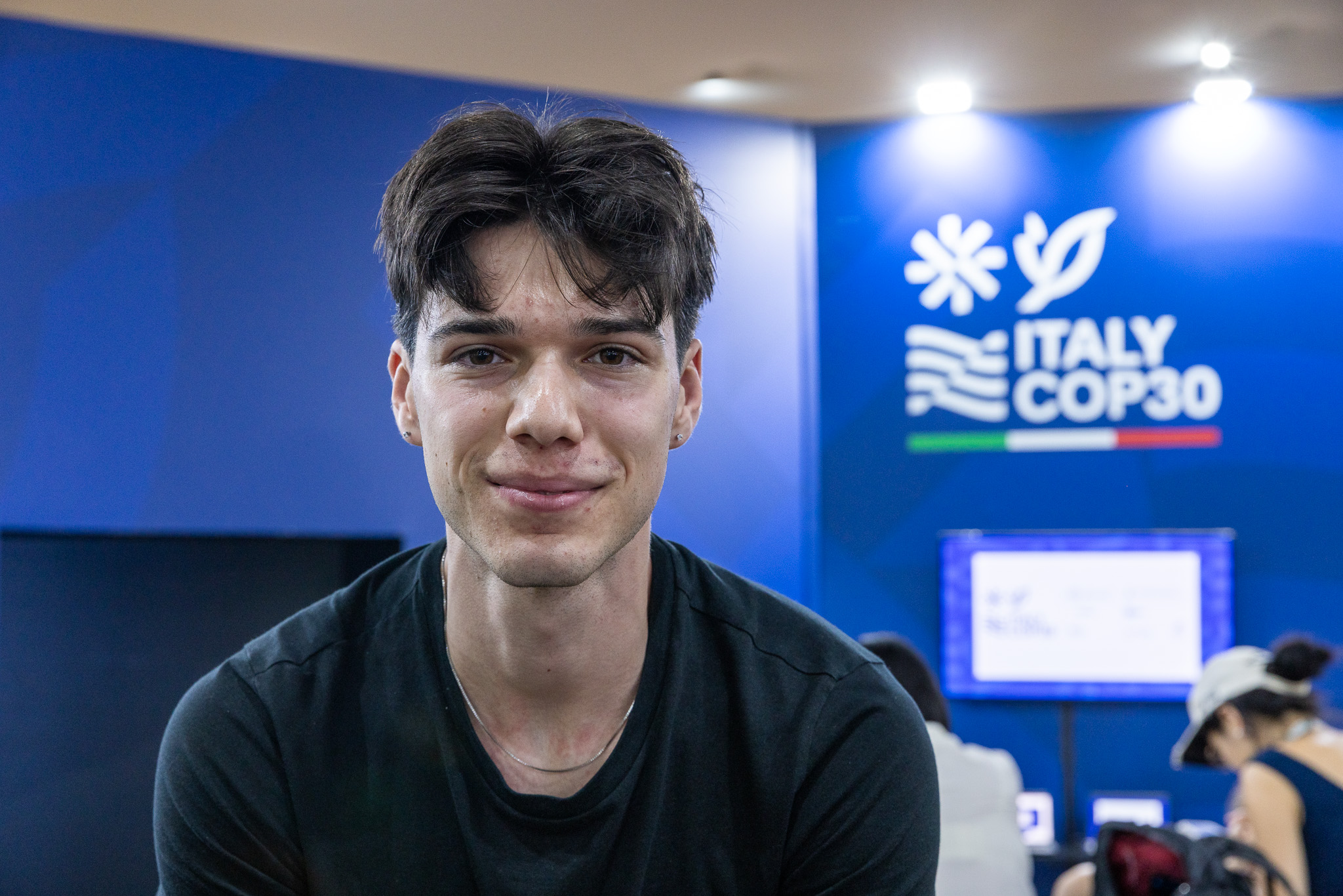 A young man in a black shirt smiles into the camera. Over his shoulder, a wall reads "Italy COP30."
