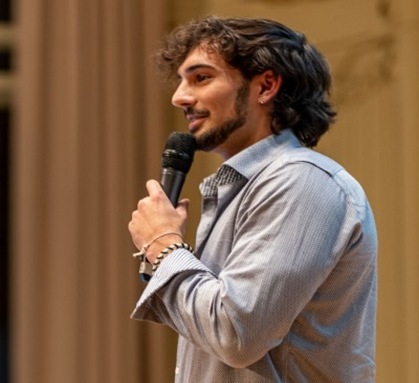Alec, dark-haired and sporting a beard, speaks into a microphone on stage.