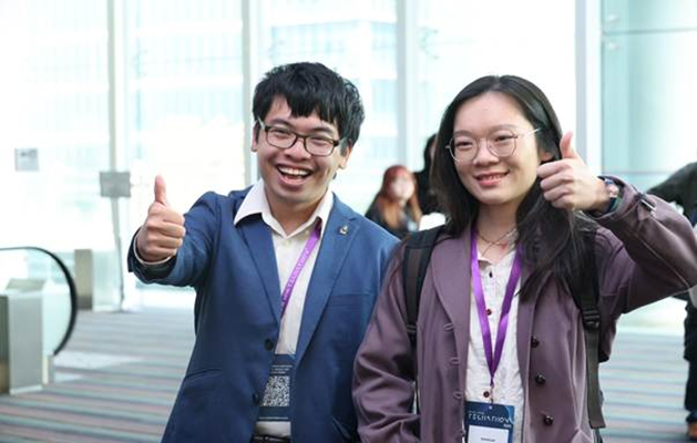 A young man and a young woman wearing glasses and event badges give thumbs-ups to the camera and smile.