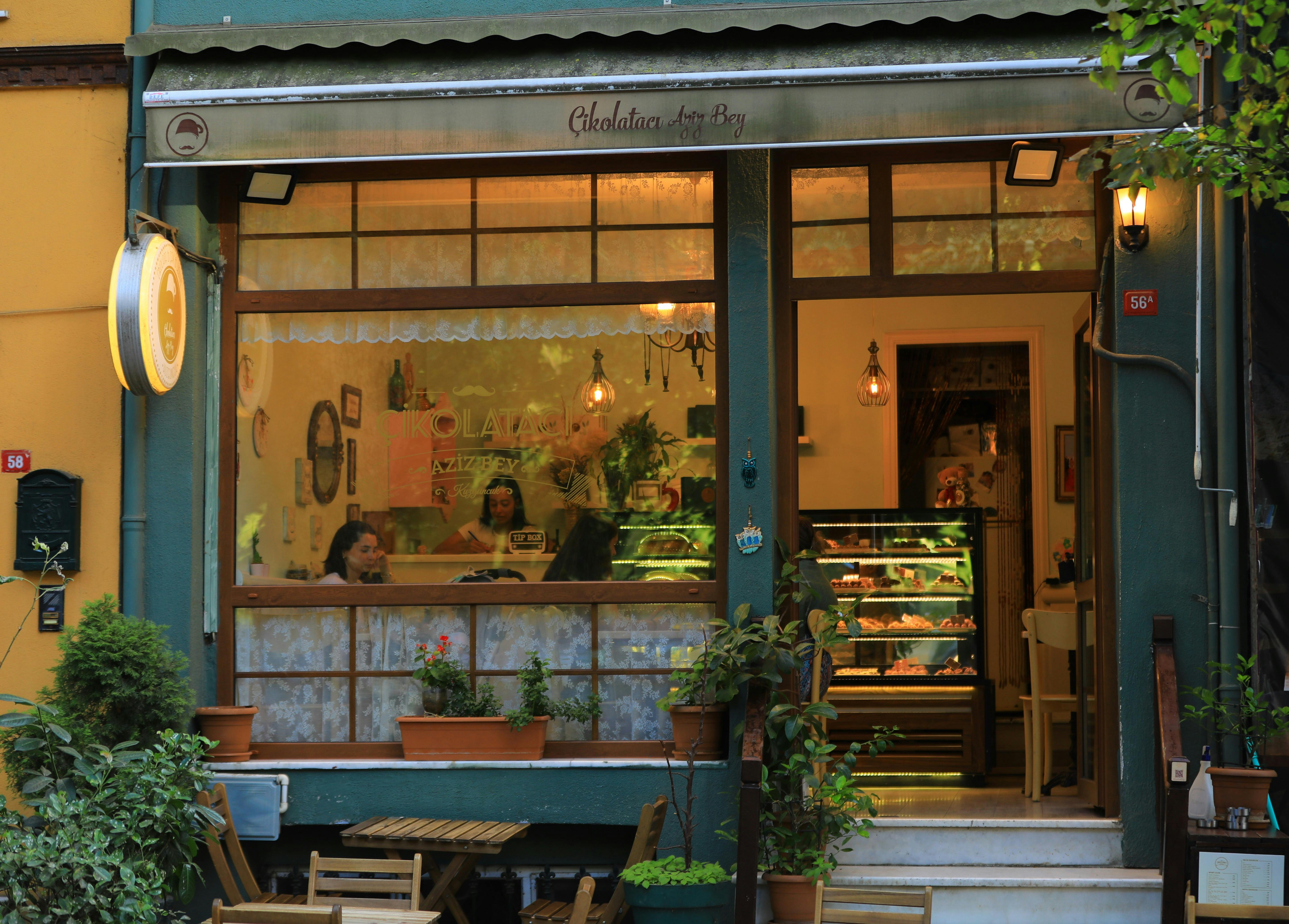 A view from the road of a cozy cafe interior. Pastries are on display, and two guests sit opposite each other at a table by the window.